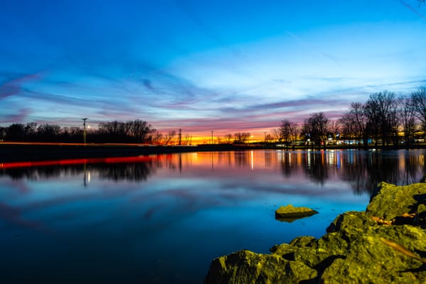 A long exposure at dusk: banded pink and orange light along the horizon, bare trees in silhouette, and still water holding the whole sky.