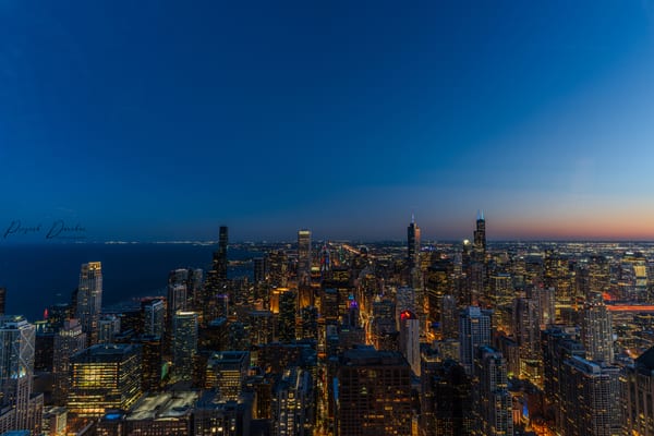 The Chicago skyline seen from above at dusk, lit windows glowing against a deep blue sky with Lake Michigan beyond.