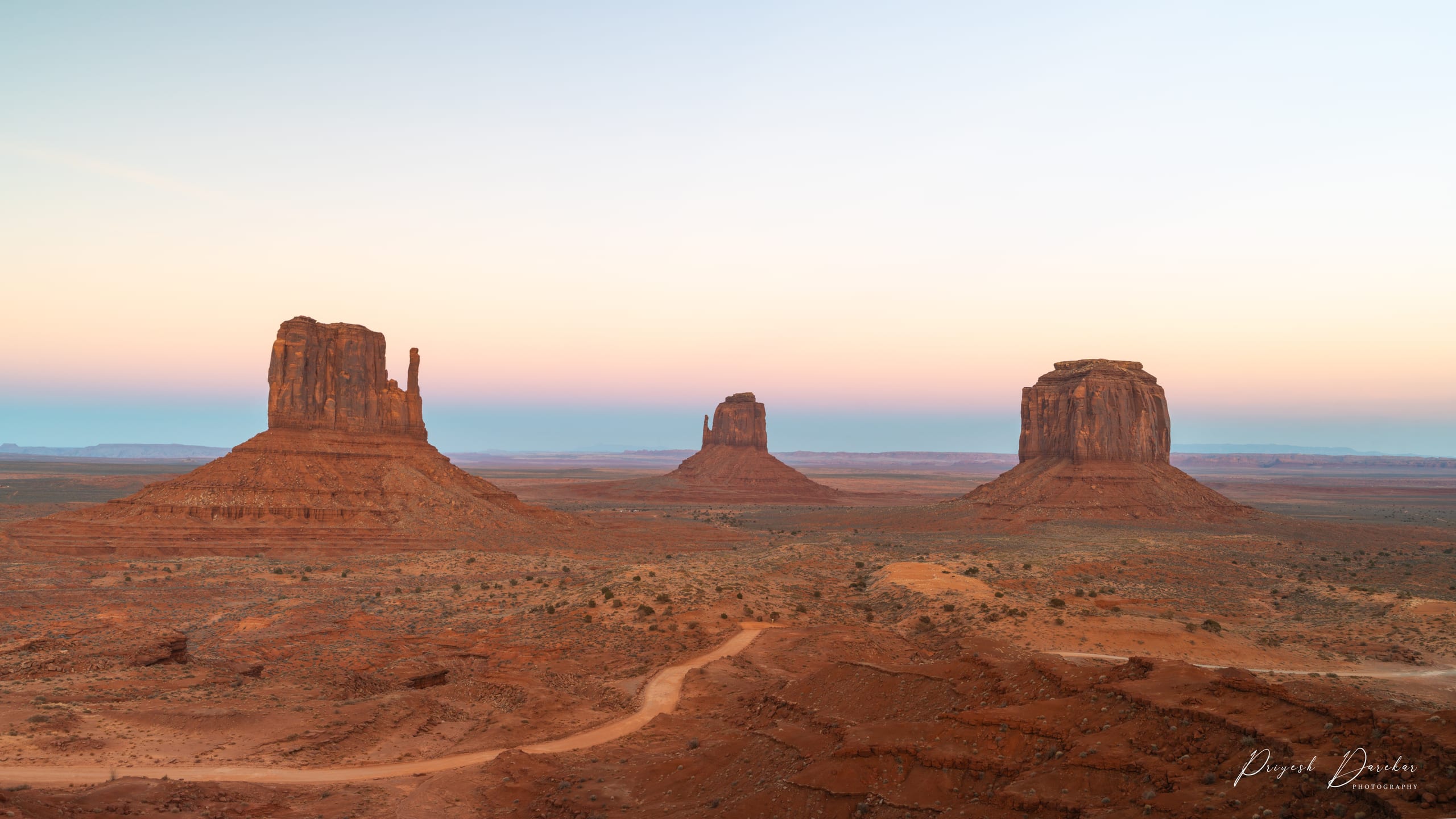 The Mittens and Merrick Butte in Monument Valley catching first light, a dirt road curving away through the foreground.