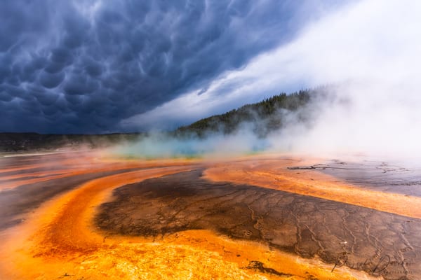Rust and amber bacterial mats sweeping across the foreground while steam lifts into a dark mammatus sky.