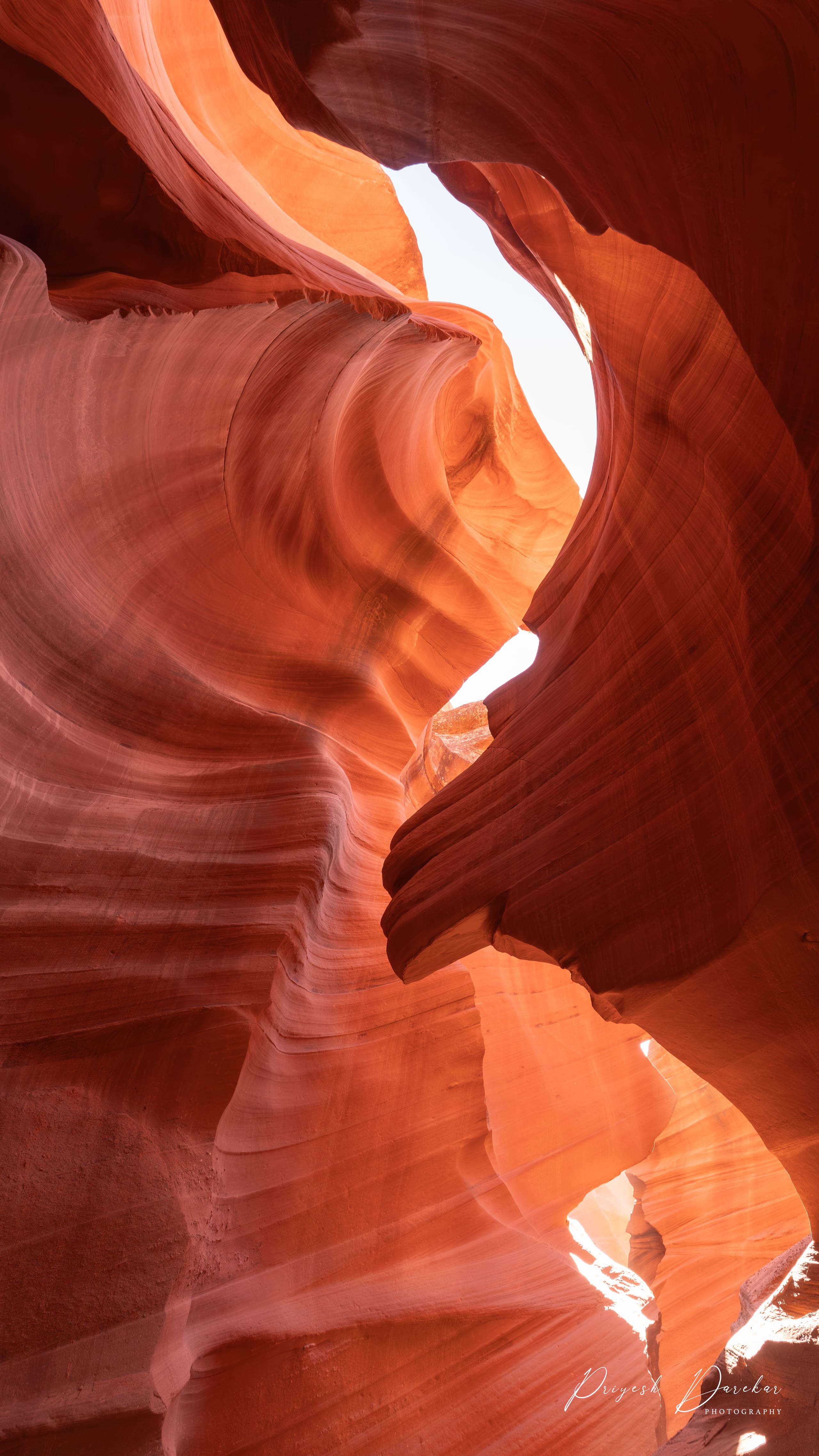 Layered orange sandstone rising on both sides of a narrow slot canyon toward a slot of white sky.