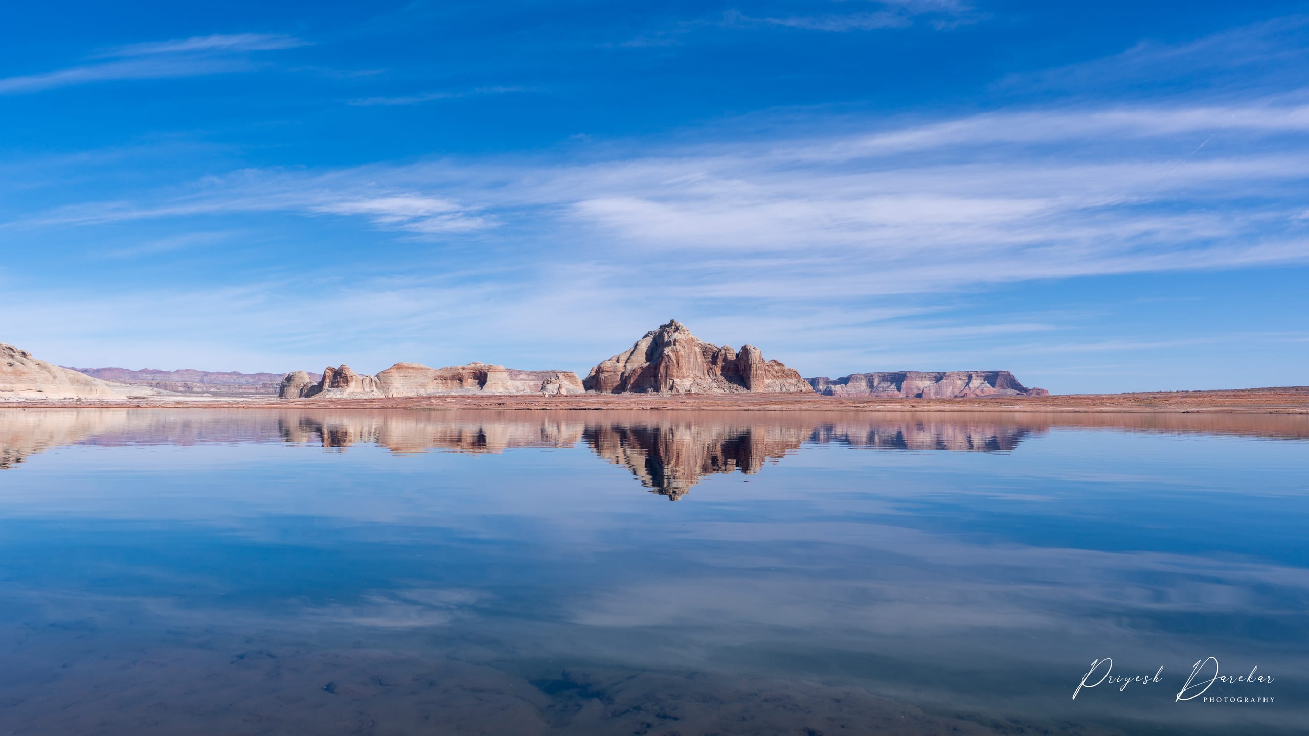 Red rock buttes mirrored in the flat blue water of Lake Powell beneath thin high cloud.